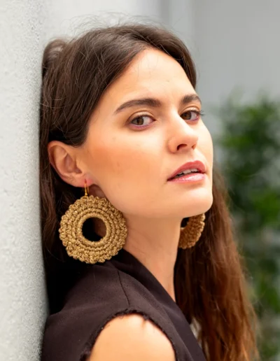 Primer plano de mujer sonriente con pendientes circulares dorados BeMeknes. Los pendientes de crochet artesanal en tono oro enmarcan su rostro, destacando la textura handmade y su brillo delicado. Joyería textil ligera para looks naturales o elegantes.